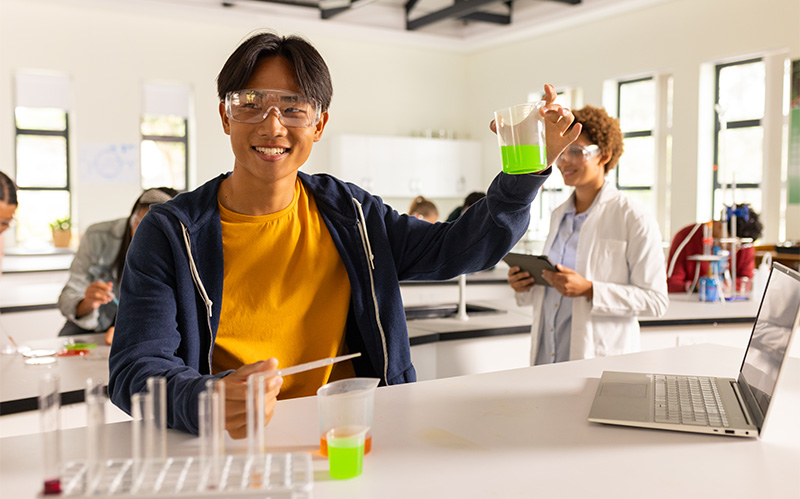 Asian student wearing safety goggles holding beaker with green liquid.
